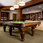 Towne Centre at Englewood photo billiards room shows pool table with pool balls, and cues. In the back ground there are seating areas with large bookshelves and a tv on the wall.